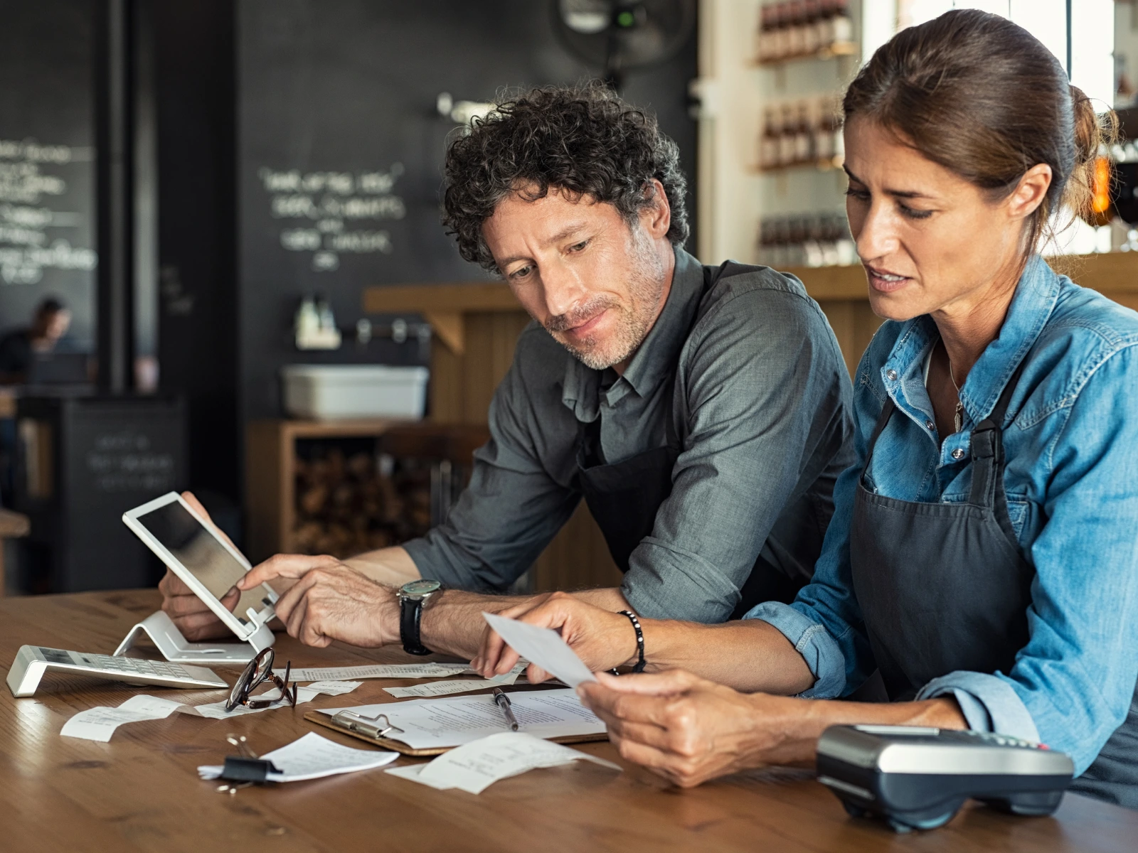 Two restaurant owners reviewing receipts and sales data together at a counter, using a tablet and calculator to track online ordering revenue.