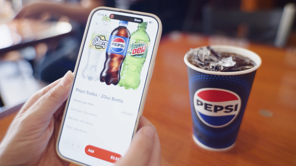 Hand holding a phone showing the ChowNow ordering app with a Pepsi soda selection screen, next to a Pepsi-branded cup of iced soda on a wooden table.
